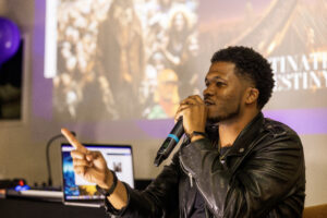 Man with microphone points to crowd with presentation behind him